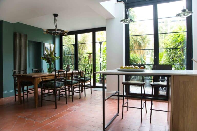 Dining area with a wooden table and chairs, a vase of flowers, large bifold doors with black frames overlooking a lush garden, and a kitchen island with stools and a bowl of fruit on top.
