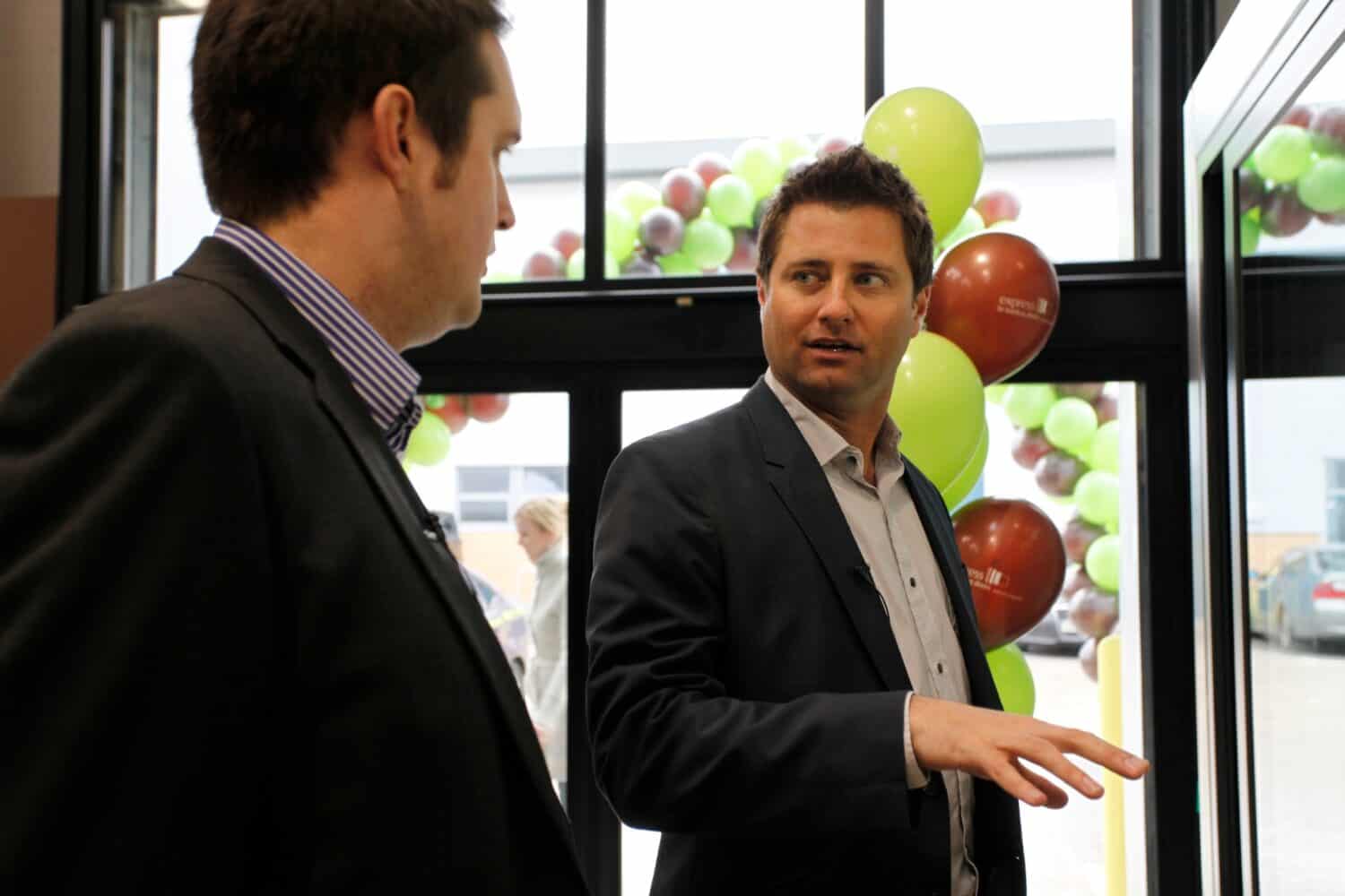 Two men in suits are talking near large windows with green and brown balloons; one gestures while the other listens. Bifold doors frame the view, with cars and a person visible outside.