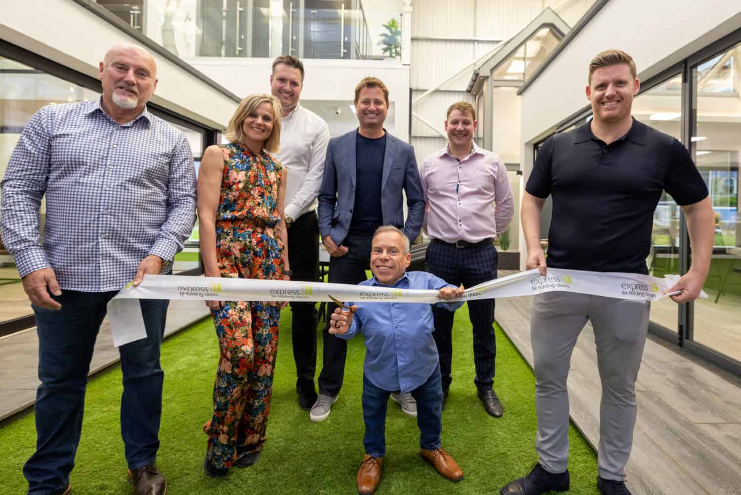 Six people smile indoors on a grass-like floor; five stand while one kneels, holding a ribbon labeled Exeed as if for a ribbon-cutting ceremony in a modern office setting featuring bespoke glass solutions.
