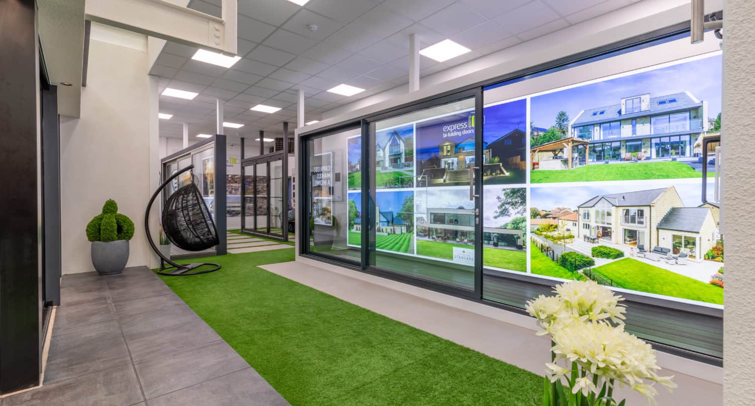 Modern indoor space with artificial grass flooring, potted plants, a black hanging chair, and a large wall display featuring photos of contemporary houses, stunning bespoke glass solutions, and stylish front doors. Ceiling lights illuminate the area.
