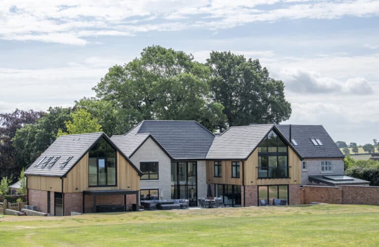 Modern two-story house with large glass windows and bespoke glass solutions, wooden and brick exterior, surrounded by green grass and trees under a partly cloudy sky.