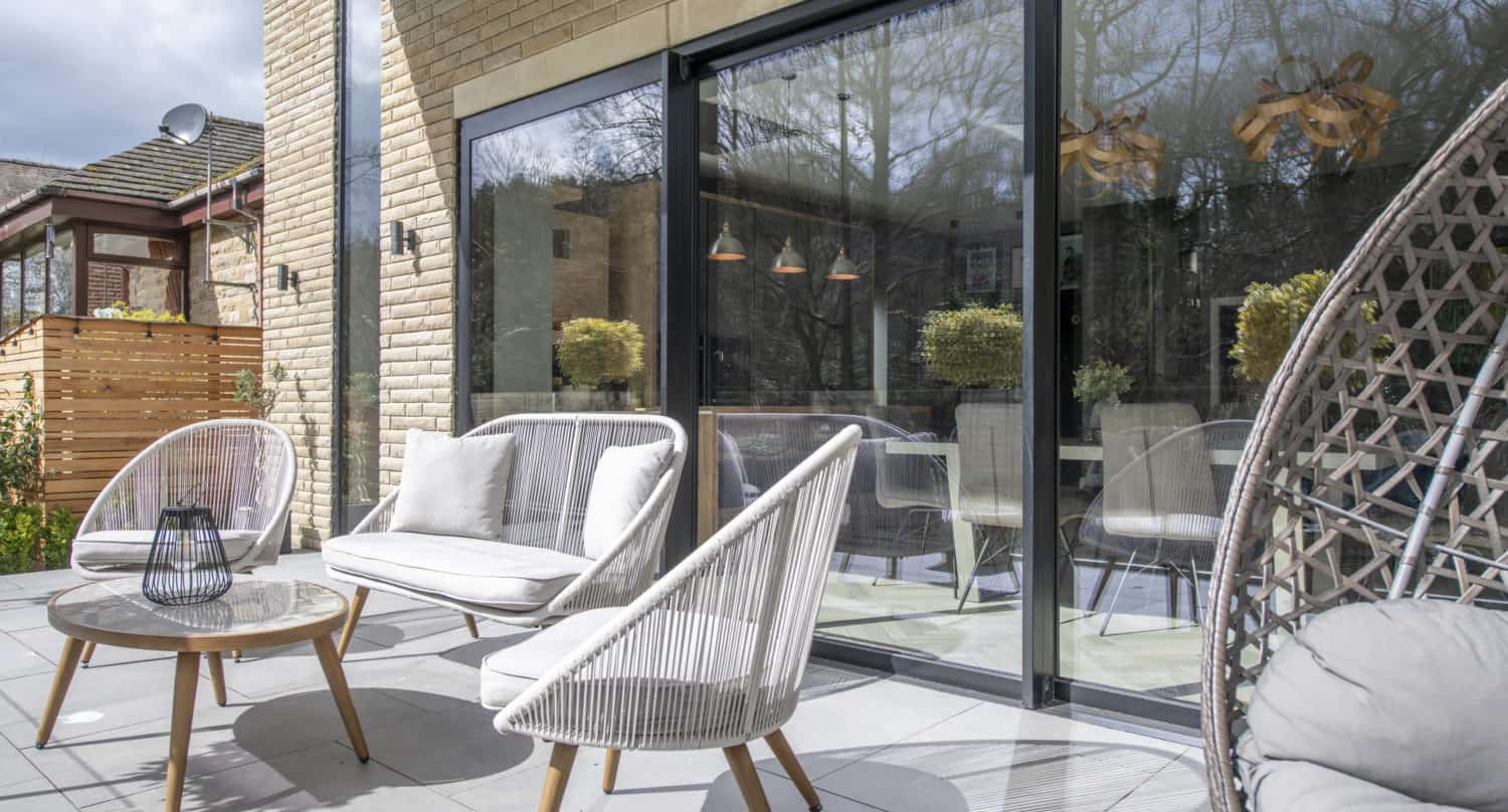 A modern outdoor patio with light gray woven chairs, a small round table, and a sofa on large tiles. Glass bifold doors reflect trees and hanging lights, seamlessly connecting the elegant patio to the interior of the house.