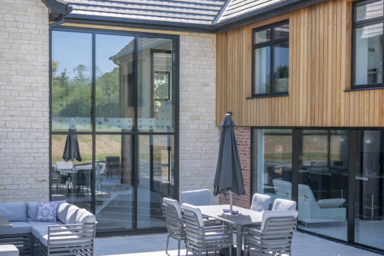 Modern house exterior with large floor-to-ceiling windows, wood and stone walls, and a patio area featuring sliding doors, a gray outdoor sofa, dining table with chairs, and closed umbrella. Trees are visible in the background.