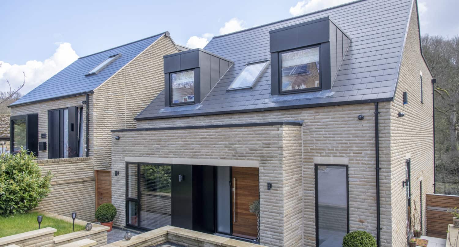 Modern two-story house with light brick exterior, large windows, black trim, and sloped dark gray roof. Features bespoke glass solutions and stylish sliding doors. Small landscaped front yard with potted plants and a driveway. Another similar building is adjacent.