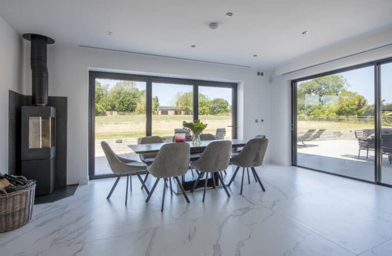 Modern dining area with a bespoke glass table and six gray chairs on white marble flooring. Large sliding glass doors open to a sunny patio with outdoor seating and green lawns. A black wood stove sits in the corner.