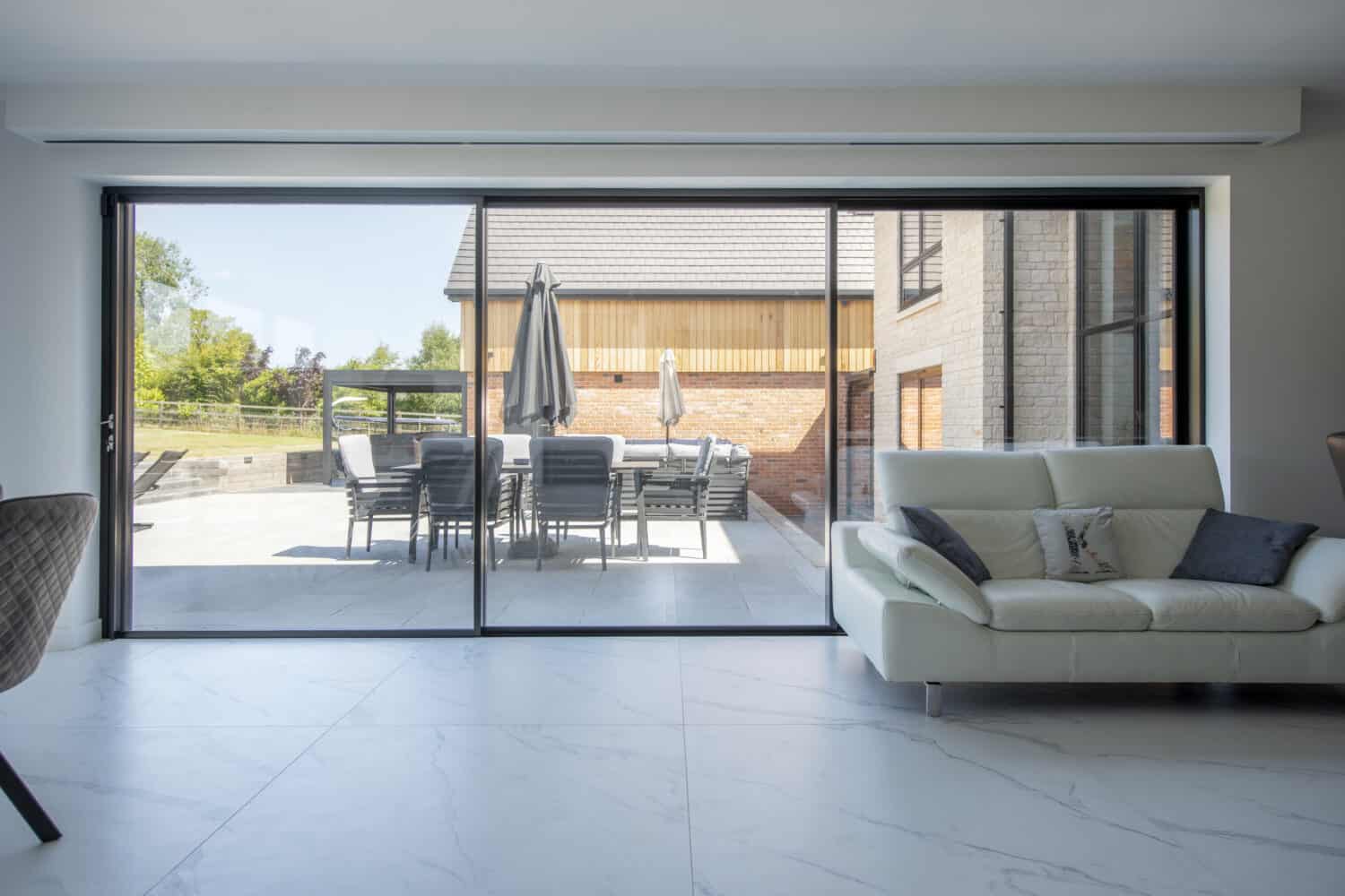 A modern living room with a white sofa and large bifold doors that open to a patio with a dining table, chairs, and closed umbrellas. The outdoor area is surrounded by trees and a contemporary house.