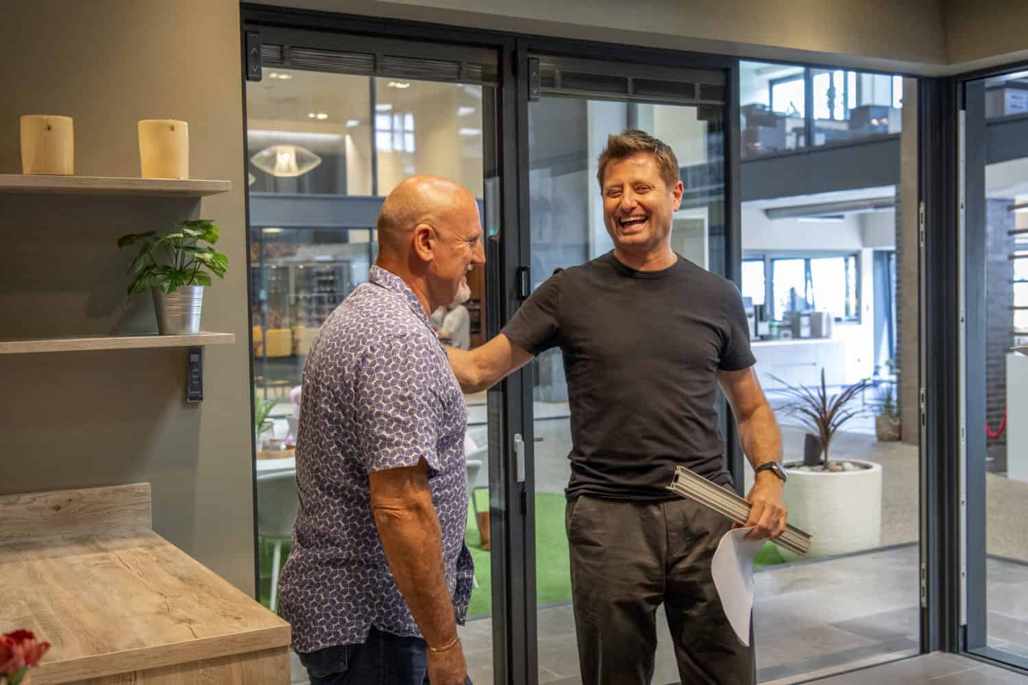 Two men stand indoors, smiling and talking. One holds documents and some strips, reaching out to the other’s shoulder. They appear to be in a modern showroom or office with glass doors and bespoke glass solutions behind them.
