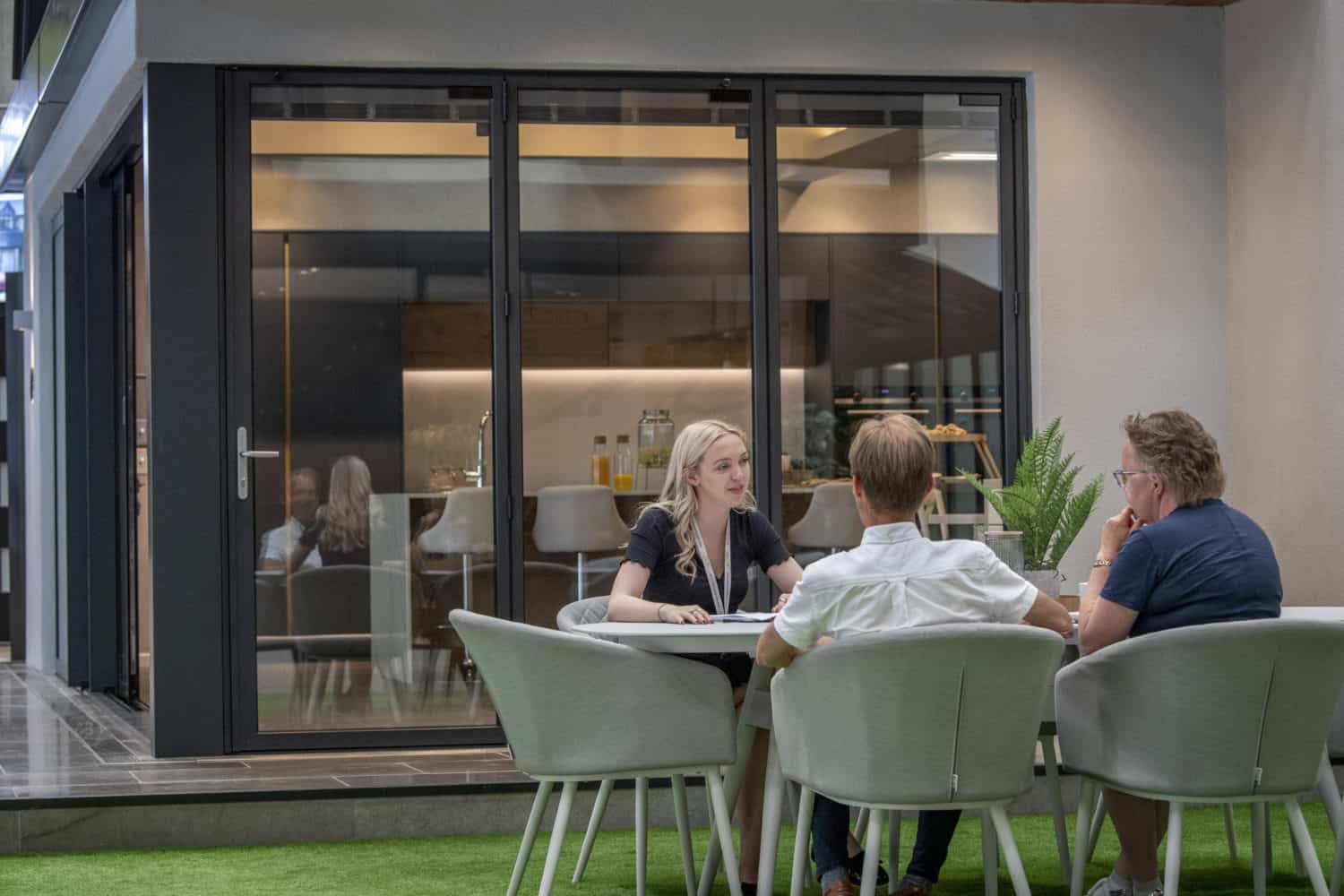 Three people sit around a table having a conversation outdoors on artificial grass, with modern sliding doors and a contemporary kitchen visible through large glass panels in the background.