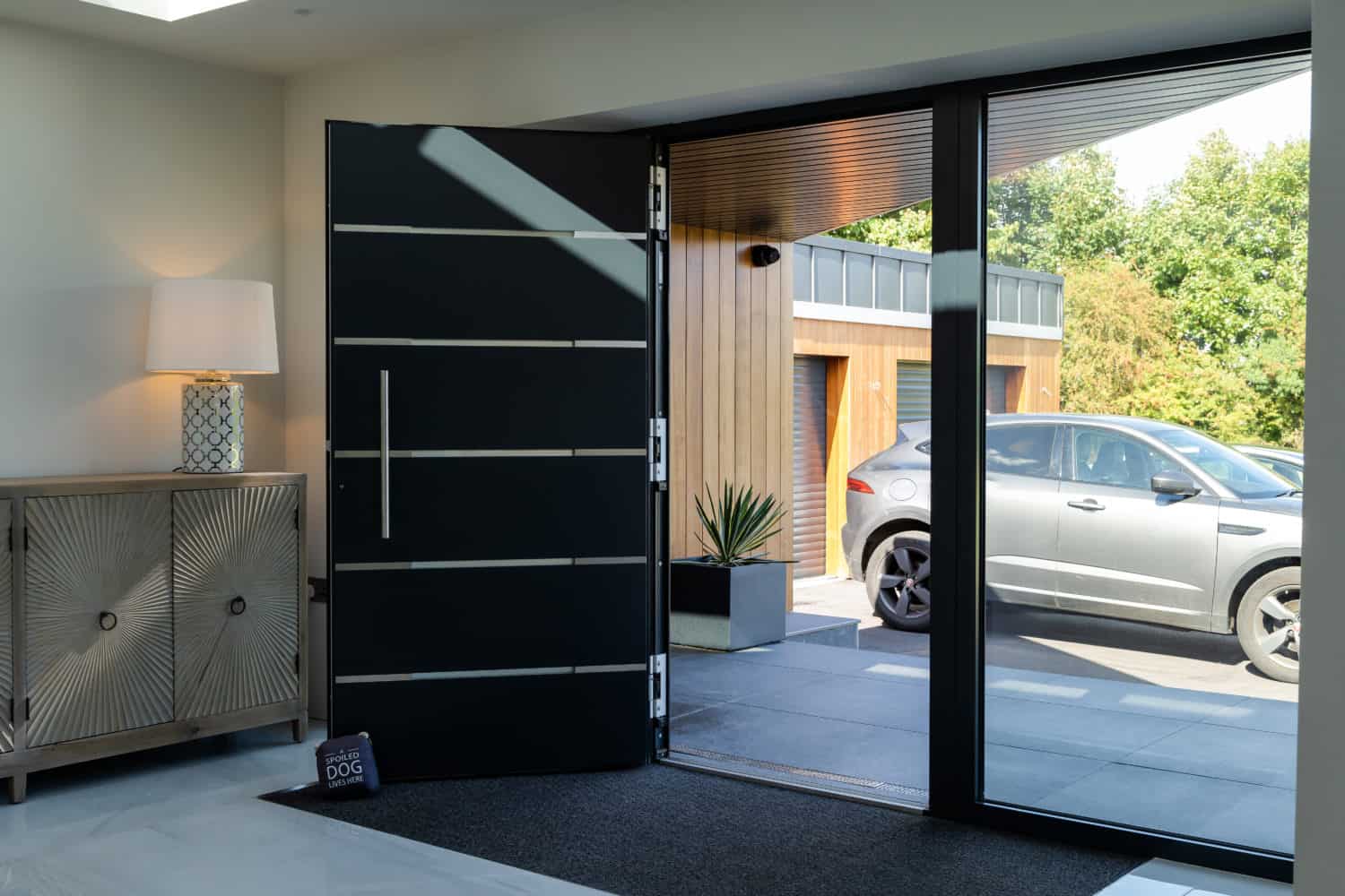 A modern front door is open, revealing a driveway with a gray car parked outside. Inside, there’s a stylish cabinet with a lamp and bifold doors nearby. Sunlight streams in through large windows onto a doormat with a sign that reads DOG.