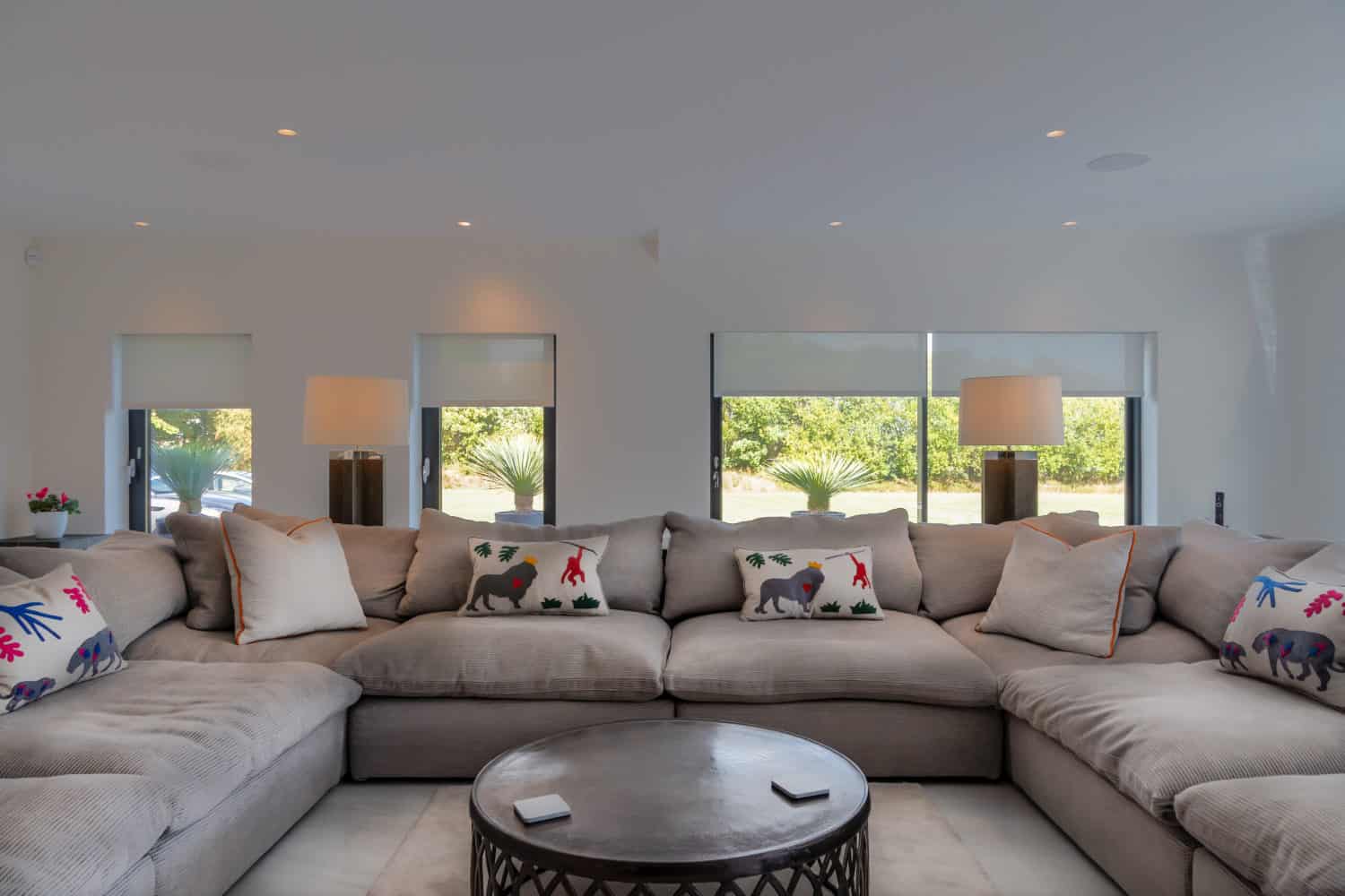 A spacious living room with a large gray sectional sofa decorated with colorful animal-themed pillows, a round coffee table in the center, and two table lamps on either side, featuring sliding doors that open to a lush green outdoor view.