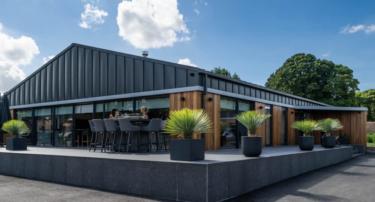 Modern building with dark metal roof and wooden accents, featuring bespoke glass solutions and bifold doors that open onto a spacious patio. Several people are seated at a high table, enjoying a sunny day under a blue sky with clouds.