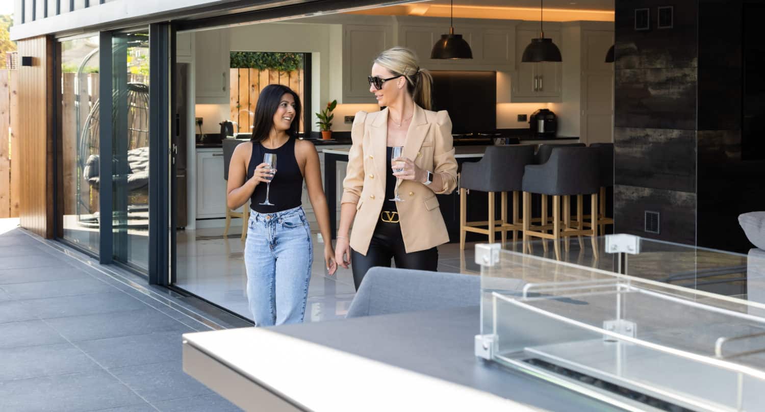 Two women, one in a tan blazer and sunglasses and the other in a black tank top and jeans, walk and talk with drinks on a modern patio beside an open kitchen, seamlessly connected by sleek sliding doors.