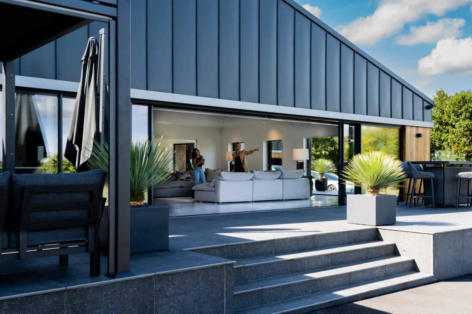 Modern house with large sliding doors opening to a terrace with gray steps, potted plants, and outdoor seating. People are sitting and talking inside a stylish, bright living room. Blue sky with clouds overhead.