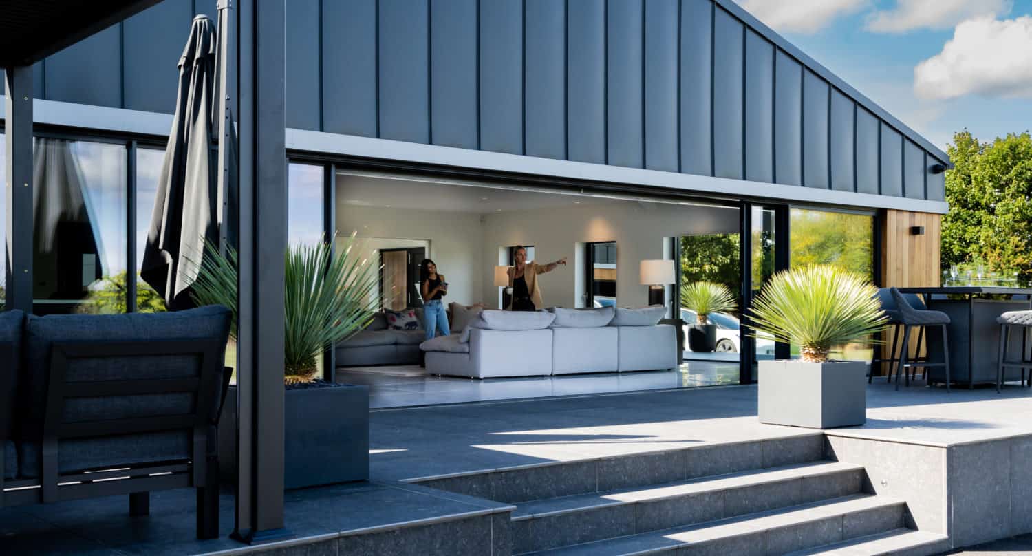 Modern house with large sliding doors opening to a terrace with gray steps, potted plants, and outdoor seating. People are sitting and talking inside a stylish, bright living room. Blue sky with clouds overhead.