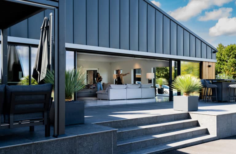 Modern house with large sliding doors opening to a terrace with gray steps, potted plants, and outdoor seating. People are sitting and talking inside a stylish, bright living room. Blue sky with clouds overhead.