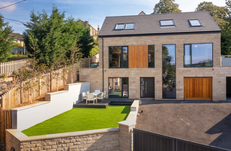 Modern two-story house with large windows, bifold doors, wood accents, and stone exterior. The yard features a walled artificial grass area with outdoor dining furniture, plus a driveway and gated entrance in the foreground.
