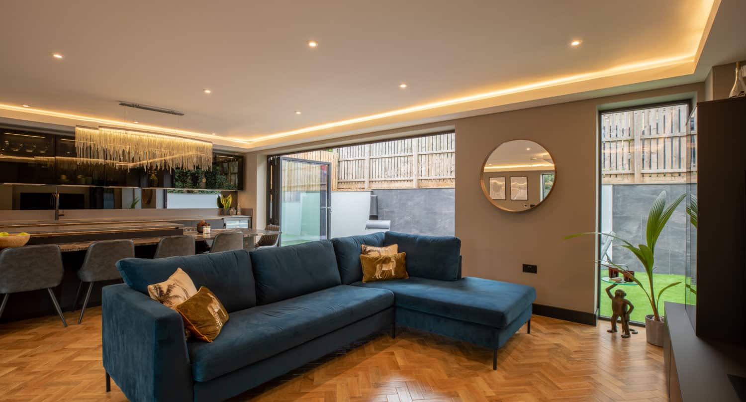 Modern living room with a blue sectional sofa, parquet flooring, open dining area, large windows, bifold doors to a garden, wall art, a round mirror, and recessed ceiling lights.