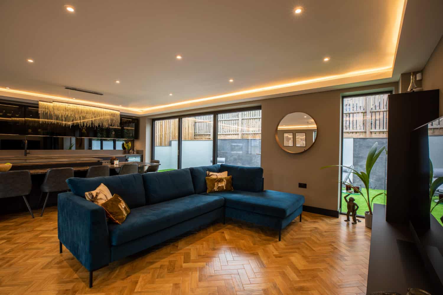 Modern living room with a blue sectional sofa, wood herringbone floor, round mirror, dining area, and large windows letting in natural light. Bespoke glass solutions like glass roofing enhance the airy feel, complemented by warm recessed lighting.