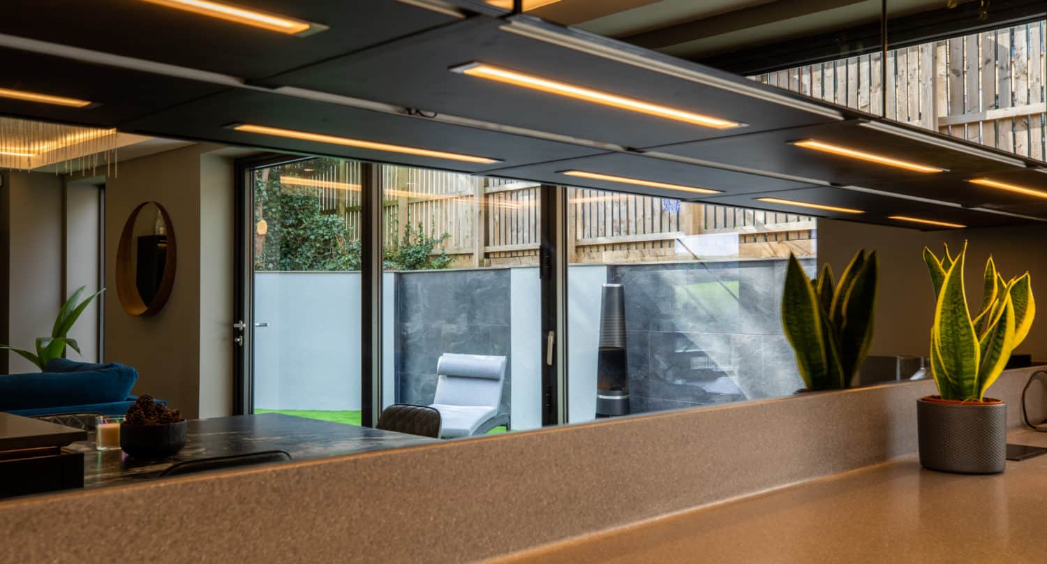 Modern kitchen with a beige countertop, indoor plants, sleek overhead lighting, and large bifold doors leading to an outdoor patio with a lounge chair and greenery outside.