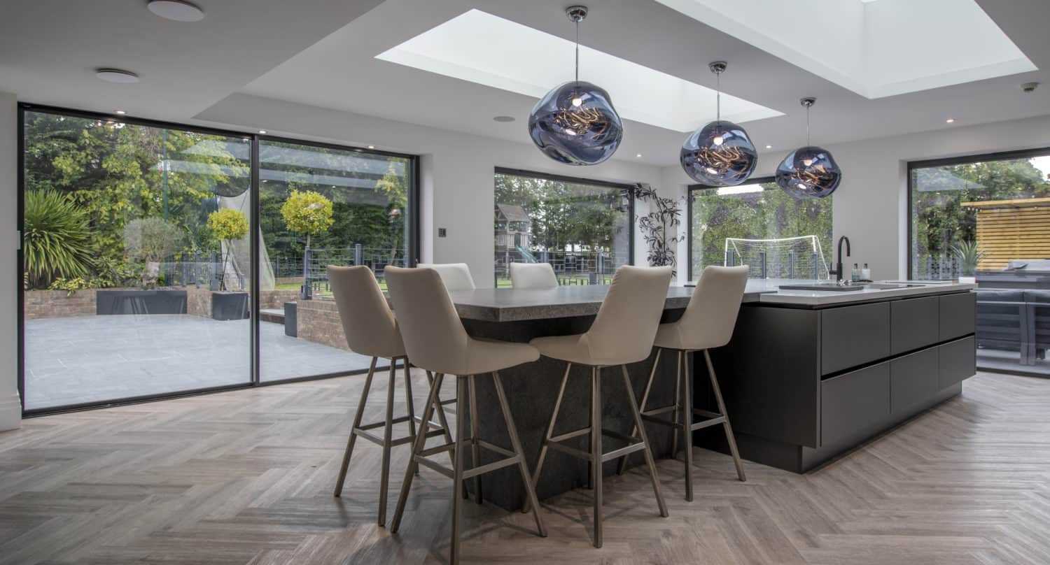 Modern kitchen with a large island, four beige bar stools, three pendant lights, skylights, and bespoke glass solutions including floor-to-ceiling sliding doors opening to a patio and garden with trees and a soccer goal.