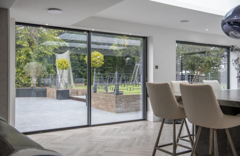 Modern dining area with tall white chairs beside sleek sliding doors, offering a view of a green garden with brick planters, outdoor seating, and trees. Light wood flooring enhances the indoor space.