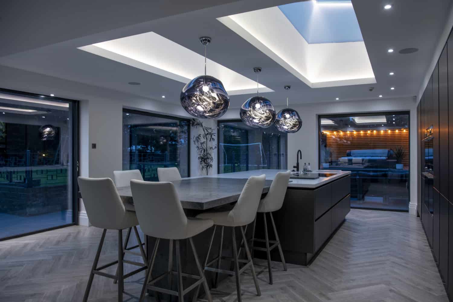 Modern kitchen with a large island, white high-backed chairs, and three artistic pendant lights. Skylights and bespoke glass solutions flood the space with natural light, with an outdoor seating area visible through bifold doors.