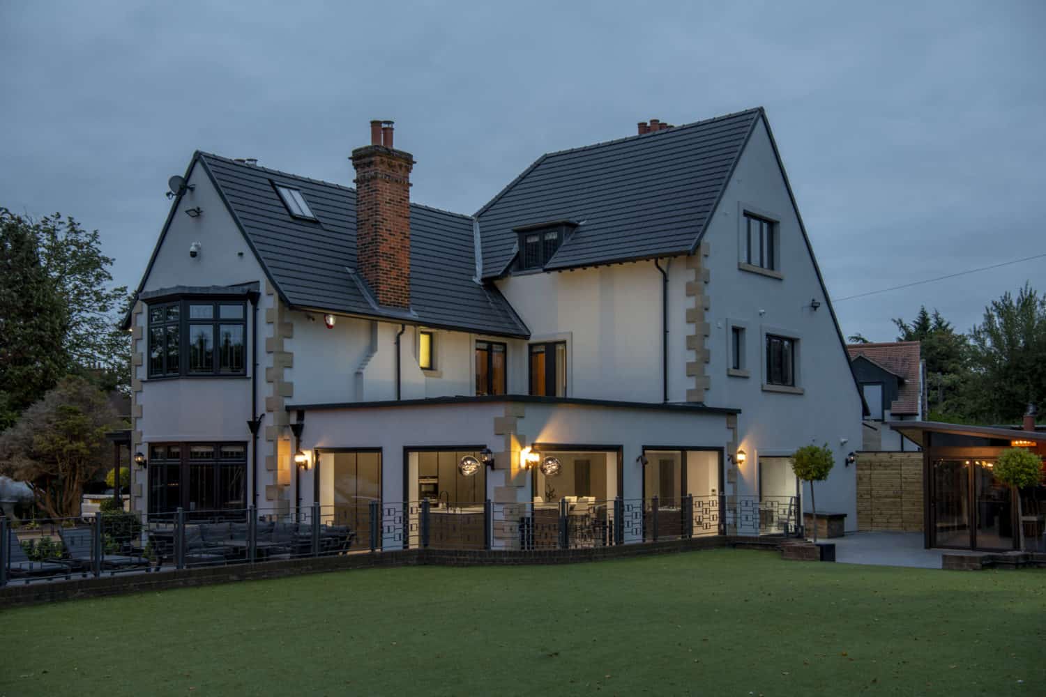 A large, modern two-story white house with black trim, lit warmly at dusk. The home features multiple large windows and sliding doors, a fenced patio area, and a well-kept lawn. Trees and outdoor lights surround the property.