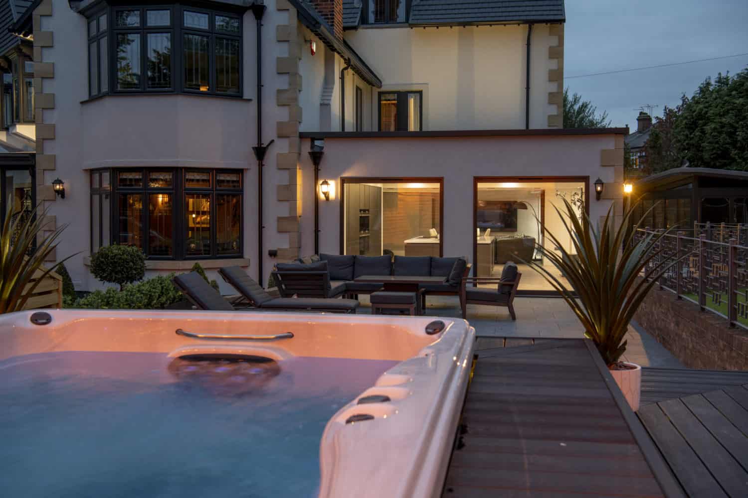 Evening view of a modern house with large windows, sliding doors opening to a patio area with outdoor seating, and a lit hot tub in the foreground surrounded by decking and plants.