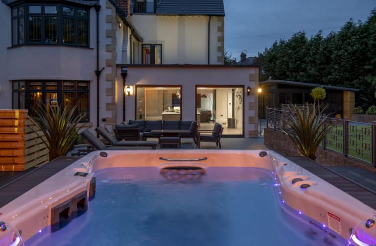 Evening view of a modern house with large windows and bespoke glass solutions, featuring an outdoor seating area centered around a lit hot tub with bubbling water. Shrubs and privacy fencing surround the patio space.