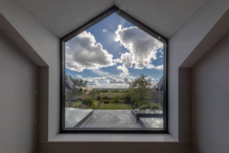 A large, house-shaped window frames a scenic view of a garden with trees and an expansive blue sky. The window’s geometric frame contrasts with the landscape outside, showcasing how bespoke glass solutions blend modern style with nature.