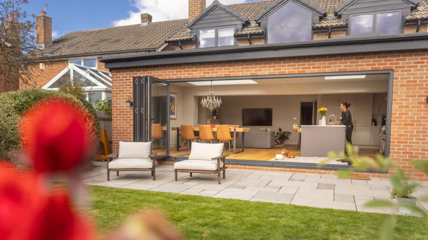 A modern brick house with large bifold doors opening to a patio and garden. Inside, a kitchen and dining area are visible. Two lounge chairs sit on the patio, and a person stands at the kitchen island.
