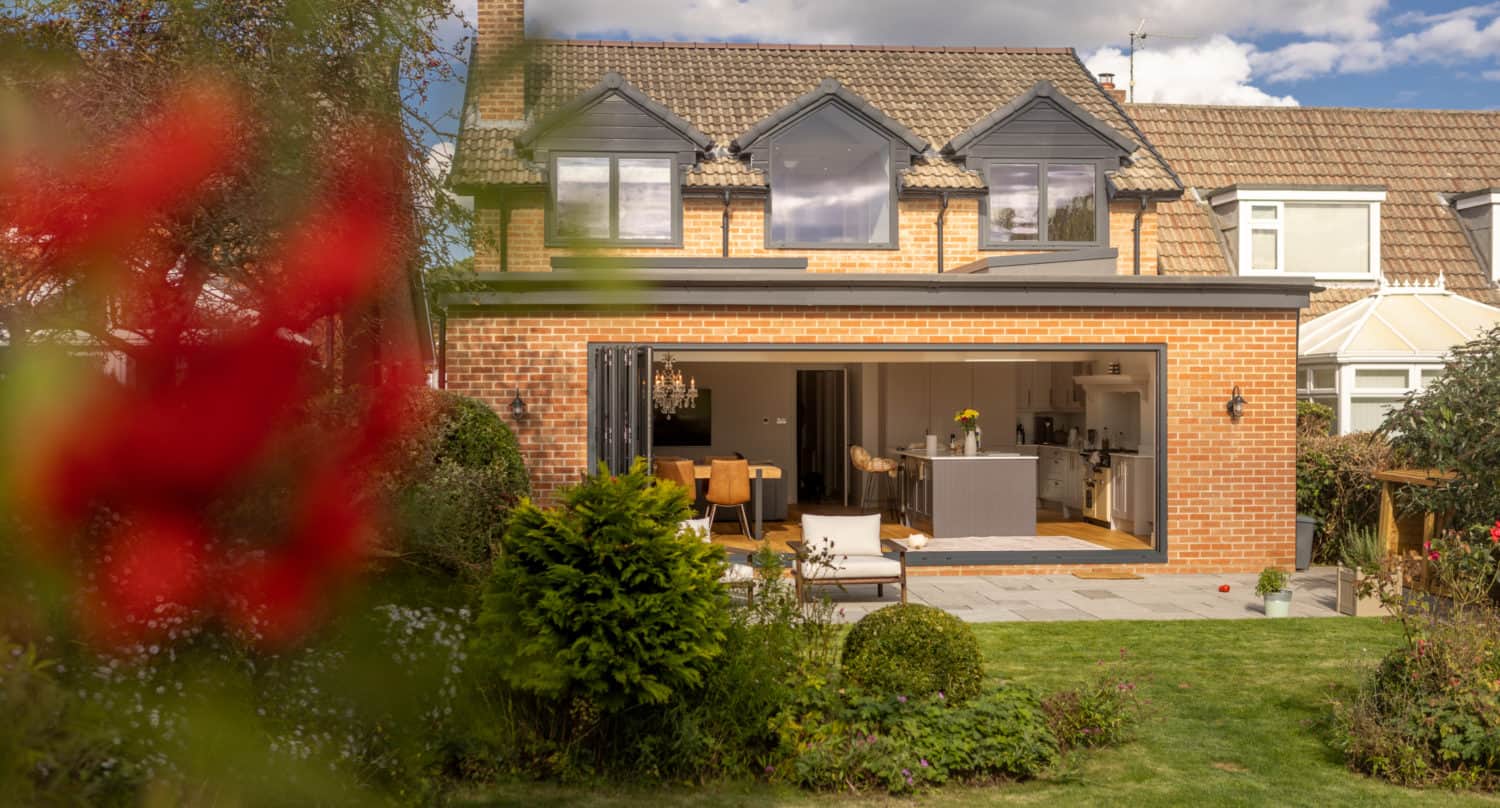A modern two-story house with an open-plan kitchen and dining area visible through large sliding glass doors and bespoke glass solutions, surrounded by a landscaped garden under a partly cloudy sky.