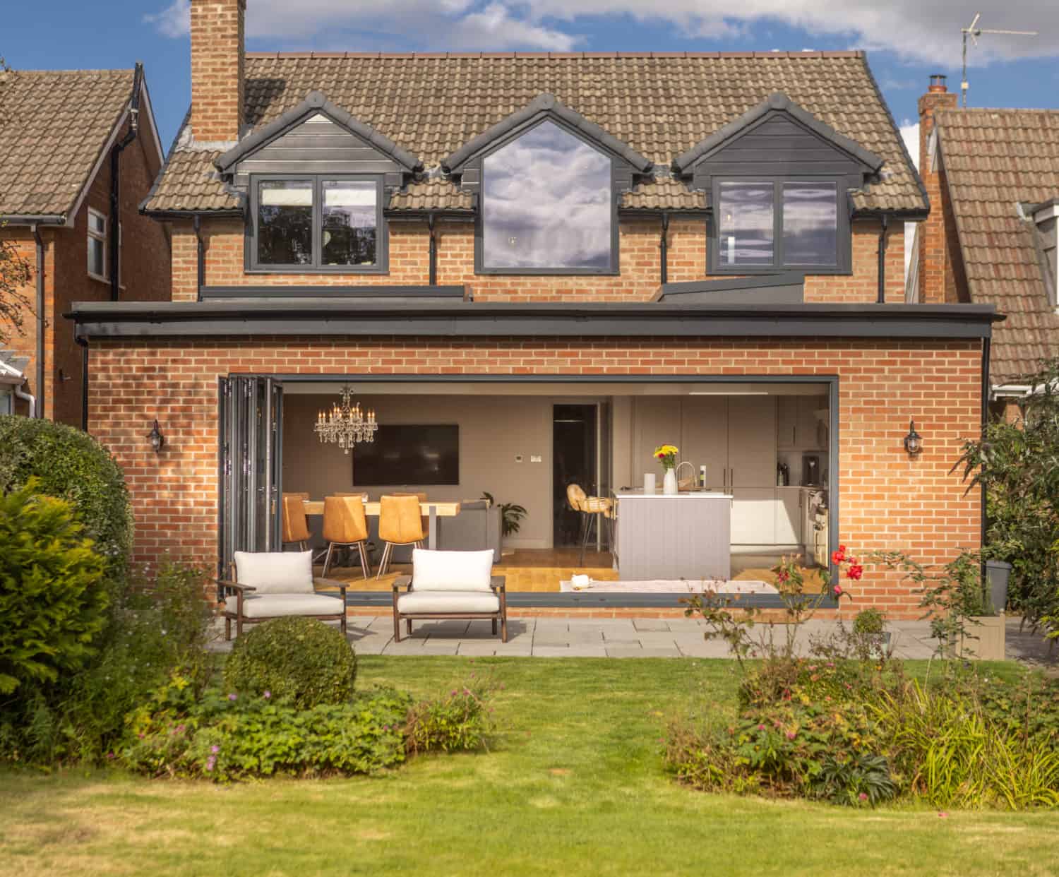 A modern brick house with bespoke glass solutions, featuring large sliding doors that fully open to reveal a spacious kitchen and dining area. The backyard offers a patio with lounge chairs, green lawn, and well-maintained shrubs and plants.
