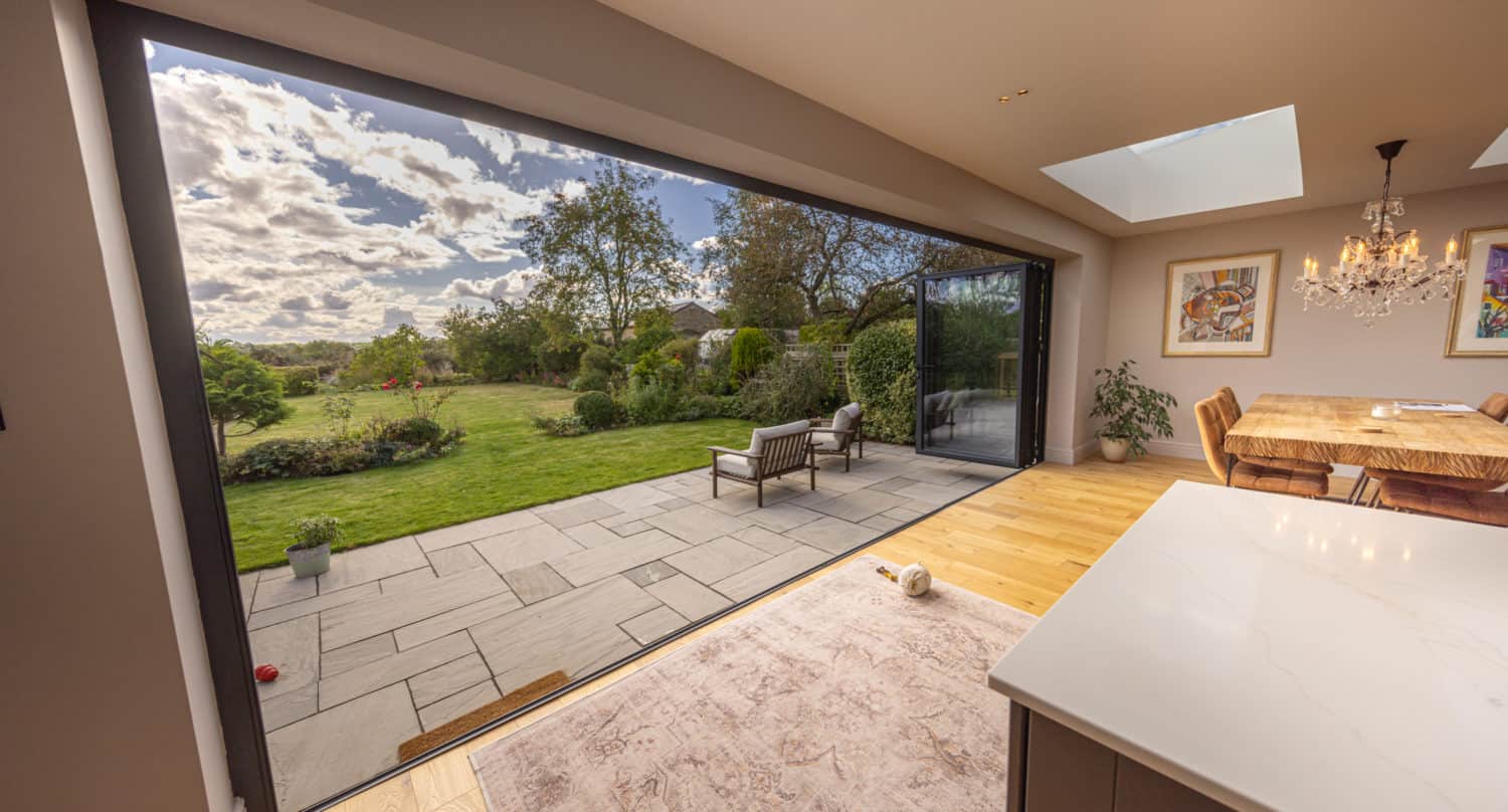A modern open-plan dining area with large sliding glass doors and skylights, leading to a sunny patio and lush green garden. Features include outdoor seating, a wooden dining table, chandelier, and stylish glass roofing.