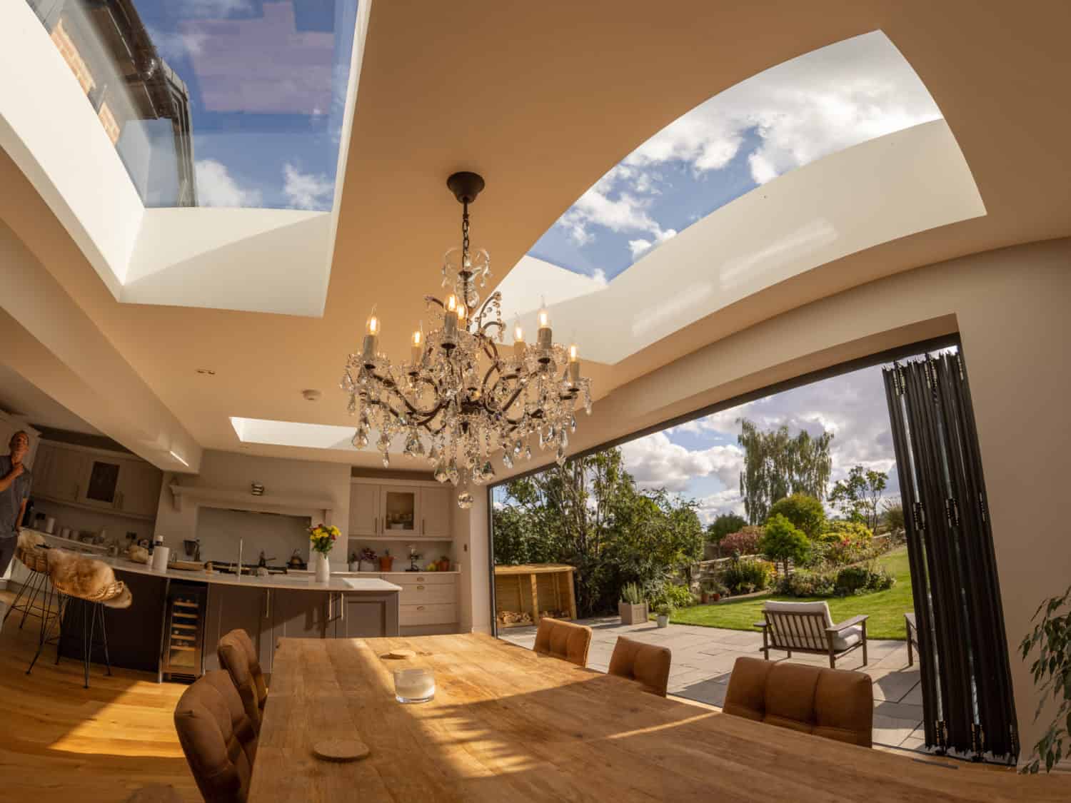 A spacious modern dining room with a wooden table, chandelier, skylights, and bespoke glass solutions like large folding glass doors opens to a sunny garden with a patio and lush greenery outside.