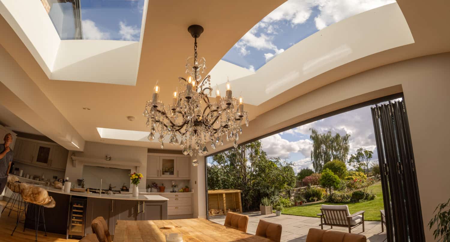 A spacious modern dining room with a wooden table, chandelier, skylights, and bespoke glass solutions like large folding glass doors opens to a sunny garden with a patio and lush greenery outside.
