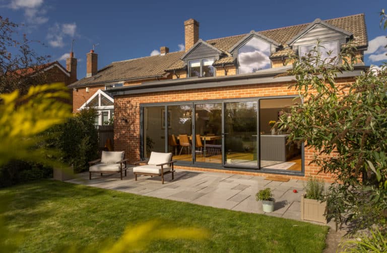 A modern brick house with large sliding glass doors opens onto a patio with two chairs. The patio, designed with bespoke glass solutions, overlooks a grassy yard with plants and trees under a blue sky with clouds.