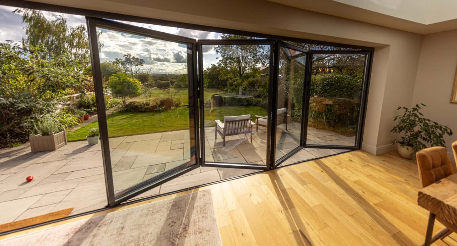 A modern living space with bespoke glass solutions, featuring open bifold doors leading to a patio and lush garden, allowing sunlight to fill the room with wooden flooring and outdoor seating visible.