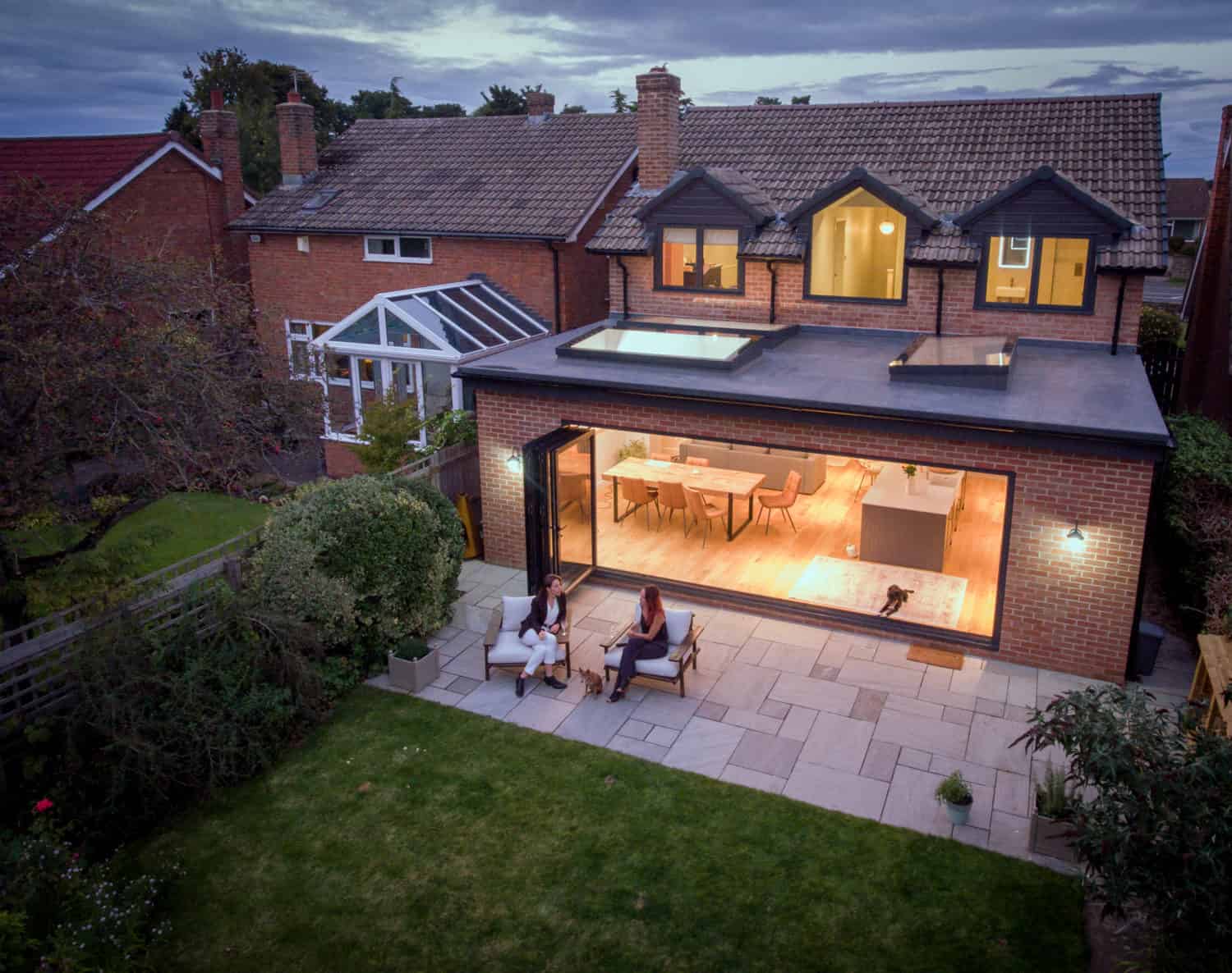 Aerial view of a modern house extension featuring bespoke glass solutions, with large glass doors opening to a patio where two people sit. The bright interior reveals a dog inside, while a lush green lawn completes the scene.