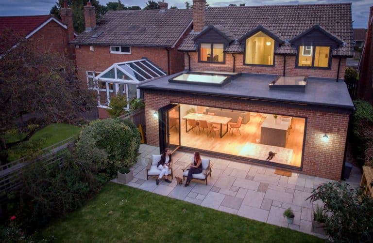 Aerial view of a modern house extension featuring bespoke glass solutions, with large glass doors opening to a patio where two people sit. The bright interior reveals a dog inside, while a lush green lawn completes the scene.