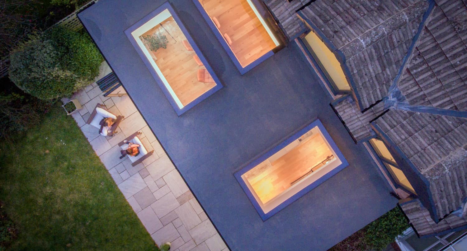 Aerial view of a modern house with bespoke glass solutions, including large skylights on a flat roof, warm lighting inside, and two people sitting at a patio table on a stone terrace beside a green lawn and surrounding garden.