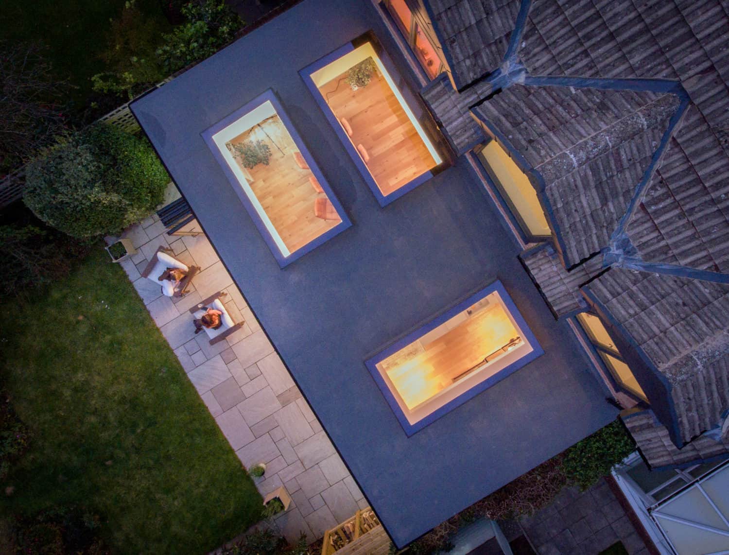 Aerial view of a modern house at dusk with bifold doors opening onto a lit patio area, two people sitting at a table, and a well-kept lawn and garden surrounding the home.
