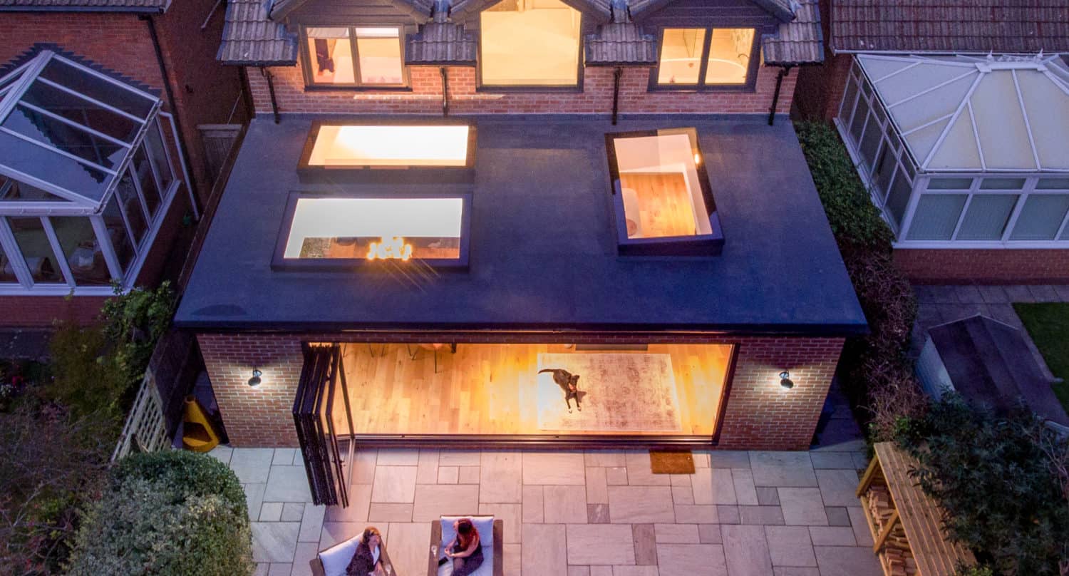 Aerial view of a modern house with large windows and sliding doors leading to a contemporary extension. Two people sit on patio chairs in the backyard, while a dog is visible inside the well-lit living room. Neighboring houses are on either side.