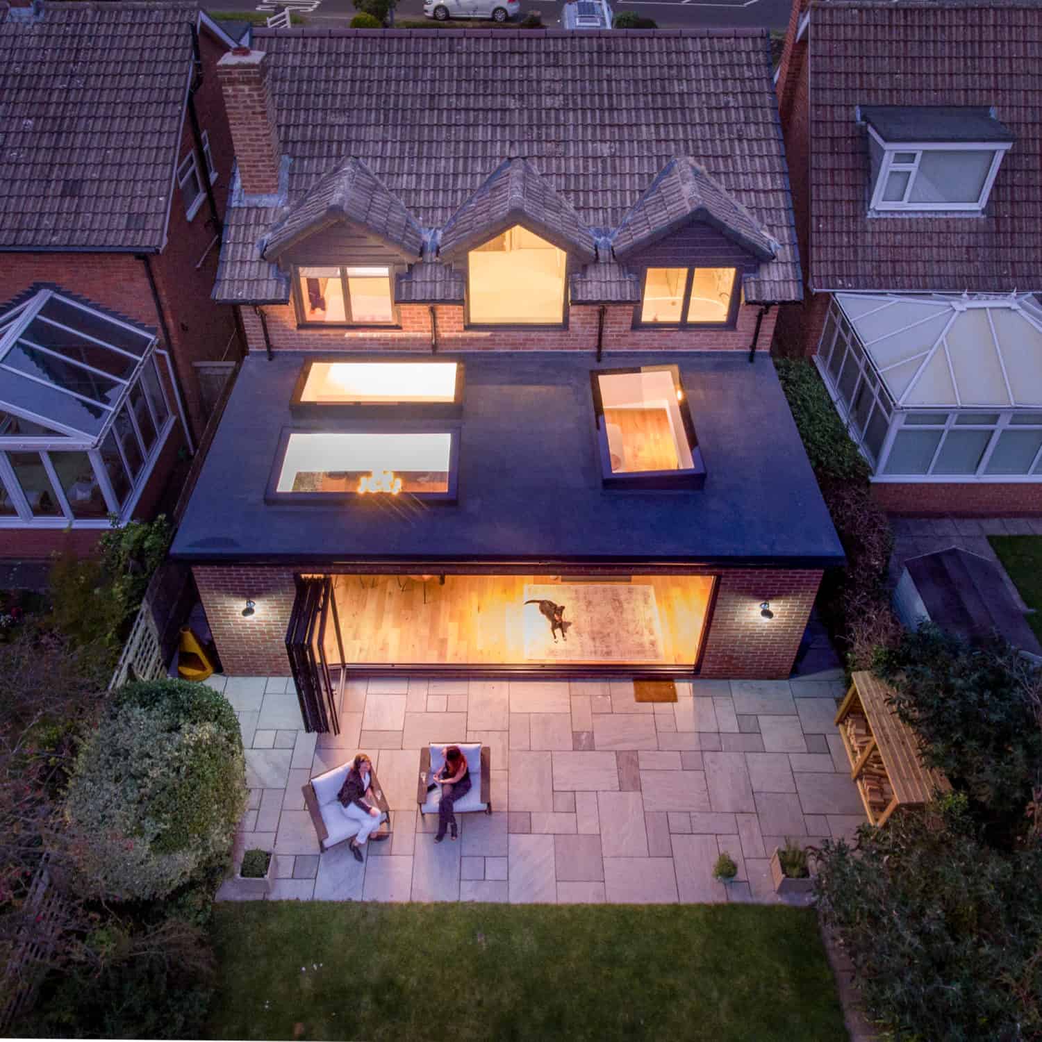 Aerial view of a modern house extension with large windows, skylights, and sleek sliding doors. Two people sit on patio chairs in the backyard, while a dog stands inside the bright interior. Evening light creates a cozy atmosphere.