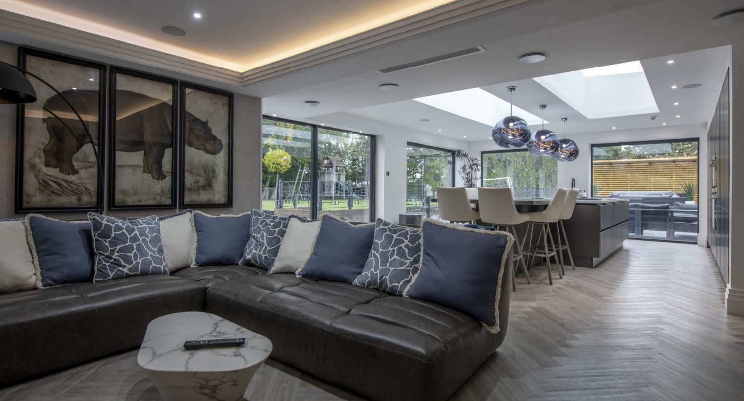 A modern living room with a large L-shaped sofa, blue and white pillows, a marble coffee table, animal artwork on the wall, and an open kitchen and dining area featuring sliding doors and large windows with skylights.