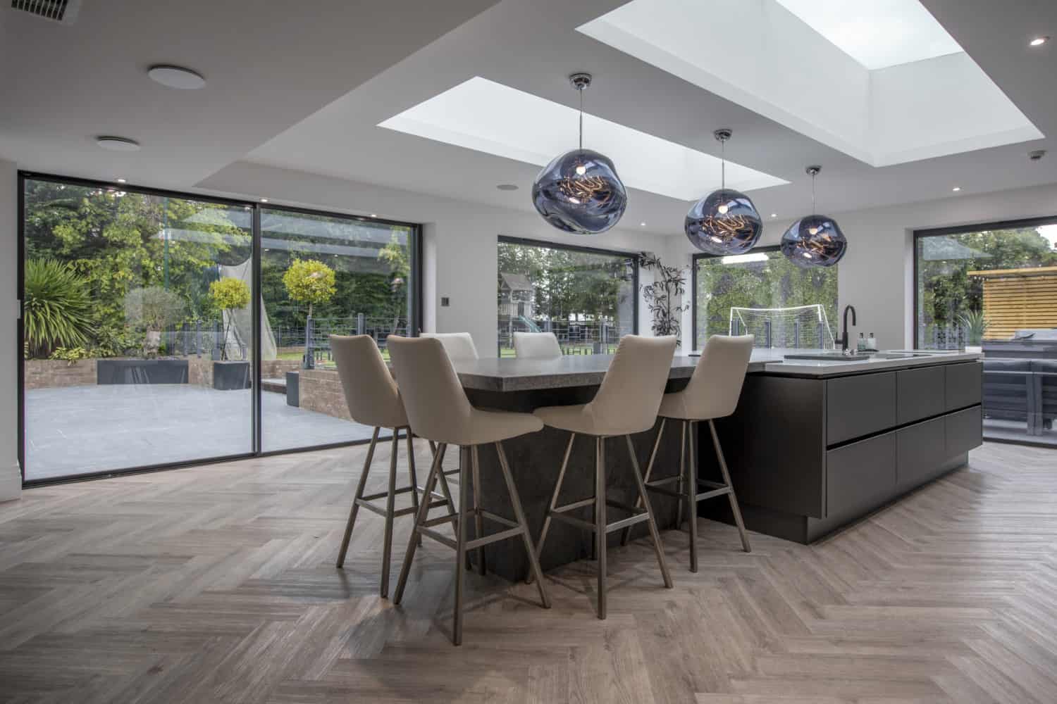 Modern kitchen with a large island, beige barstools, three pendant lights, skylights with glass roofing, and floor-to-ceiling bifold doors opening to a spacious patio and garden with greenery and outdoor furniture.