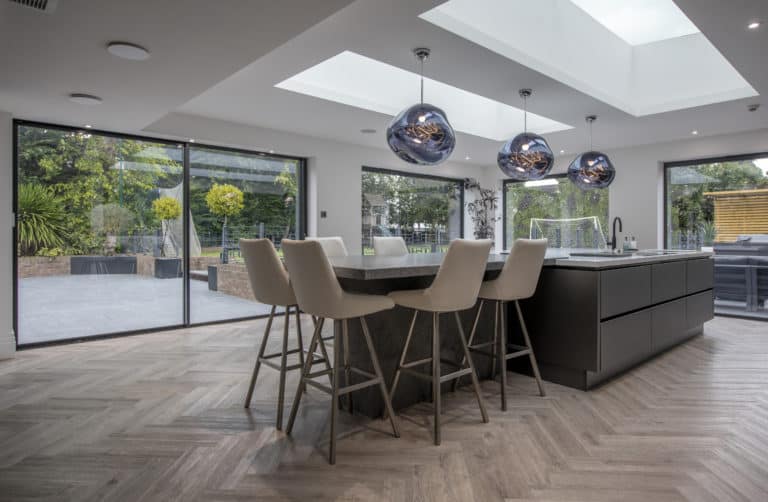 Modern kitchen with a large island, beige barstools, three pendant lights, skylights with glass roofing, and floor-to-ceiling bifold doors opening to a spacious patio and garden with greenery and outdoor furniture.