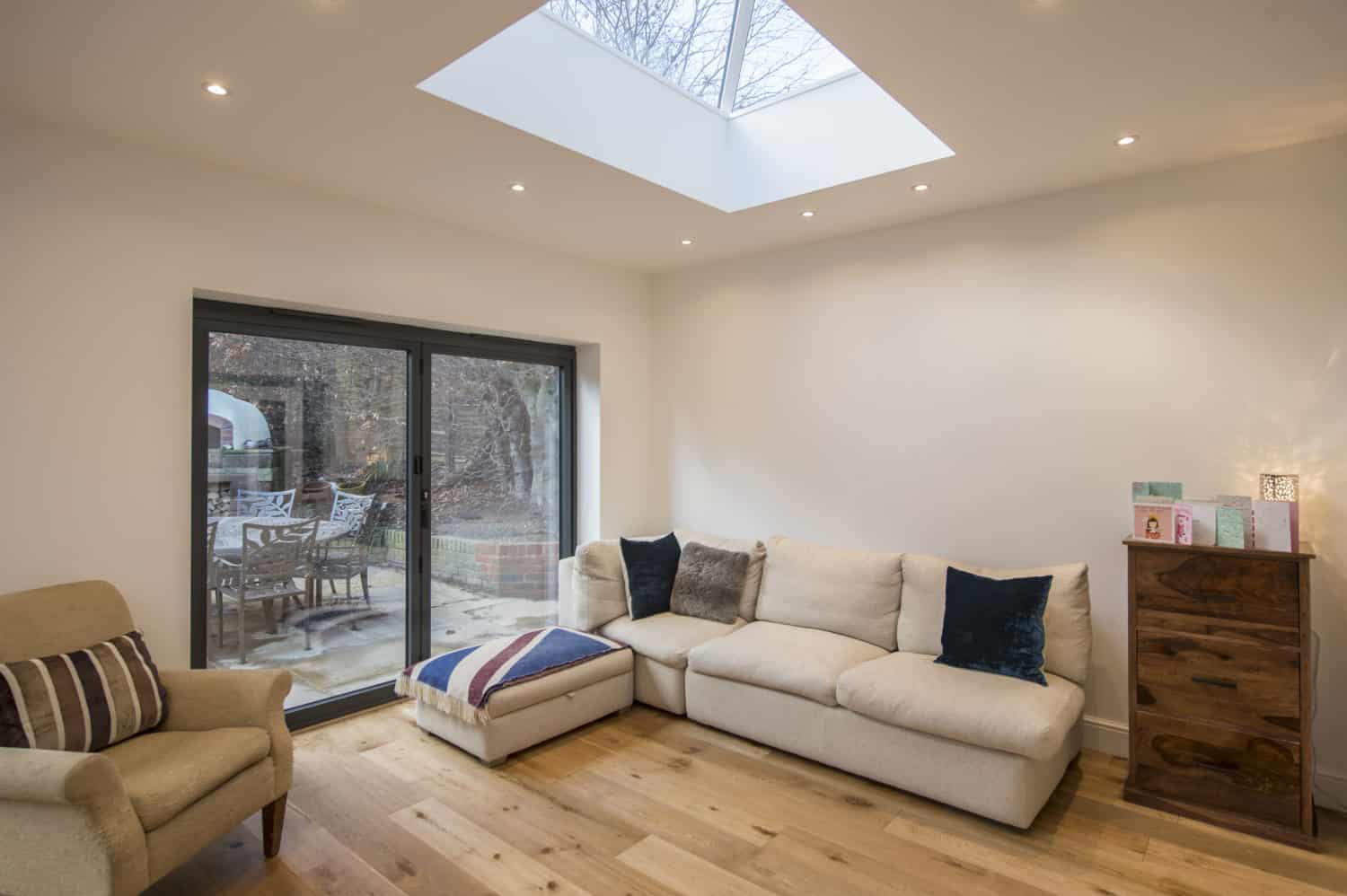 A bright living room with a skylight, light wood floor, cream sectional sofa with pillows, an armchair, and a wooden chest of drawers. Large bespoke glass bifold doors open to a patio with outdoor seating and trees visible outside.