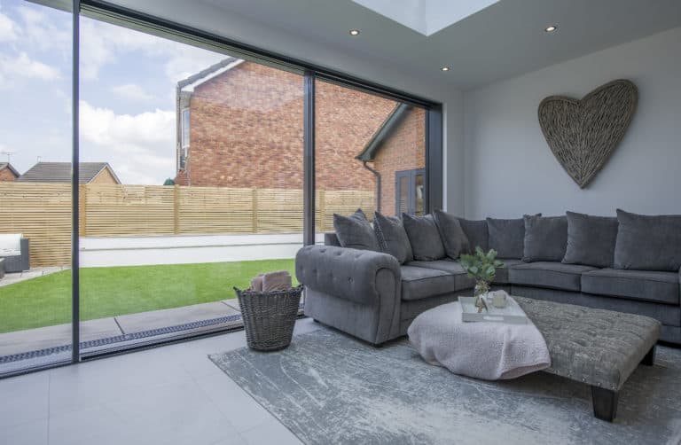 Modern living room with large bespoke sliding glass doors, gray sectional sofa, heart-shaped wall decor, and a coffee table on a gray rug overlooking a neat backyard with artificial grass and a wooden fence.