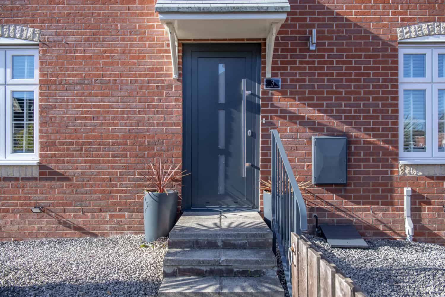 A modern dark gray front door with a metal handle, set in a red brick house featuring two windows, potted plants, and a mailbox. The entrance is enhanced by concrete steps and complemented by stylish bifold doors nearby.